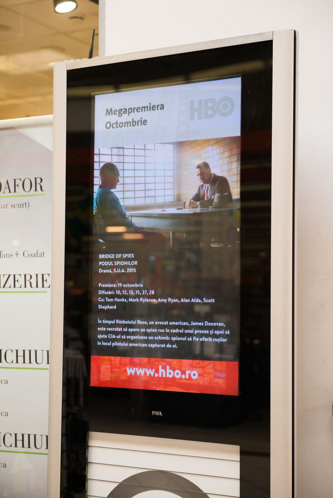 A tall totem screen fills the frame with a full-height film-premiere promo beside a shop window.