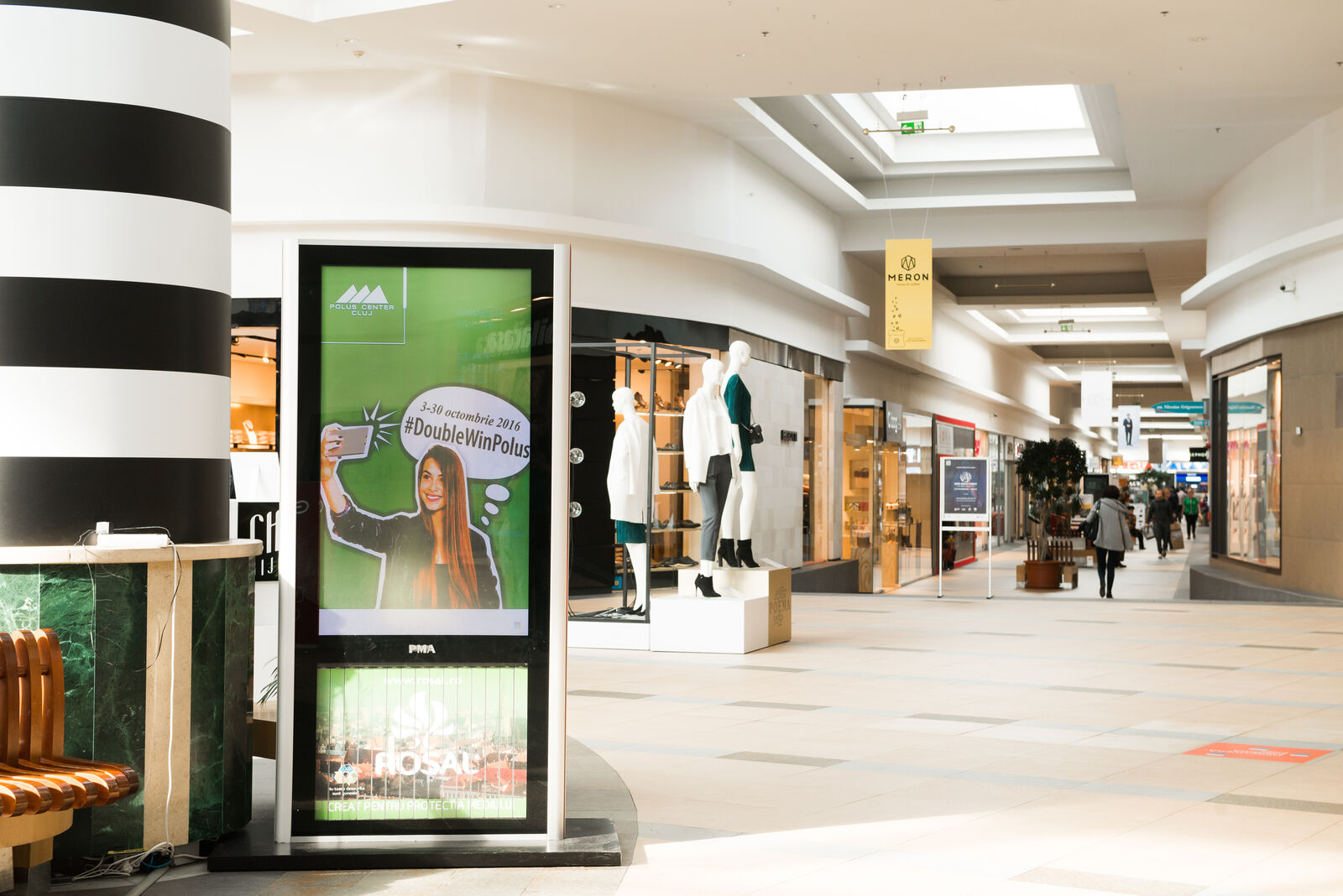 A slim totem stands at a store entrance in a bright mall concourse, running a promotional campaign.