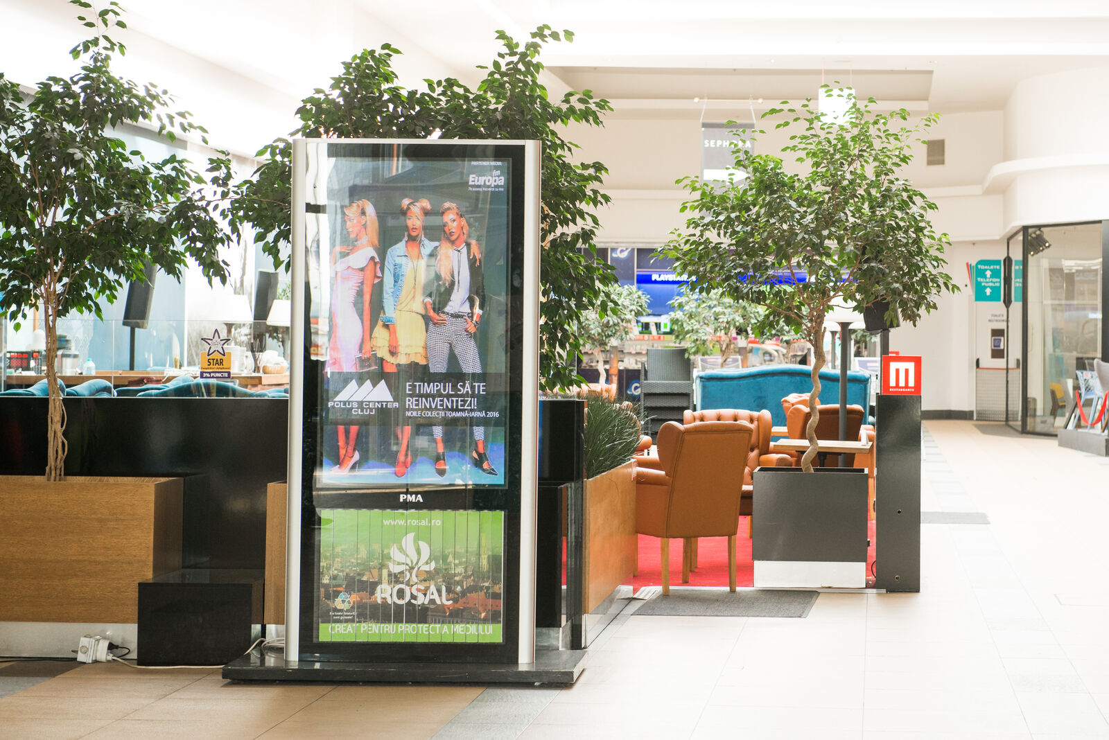 A freestanding totem anchors a plant-framed lounge in an upmarket mall, playing a seasonal fashion loop.