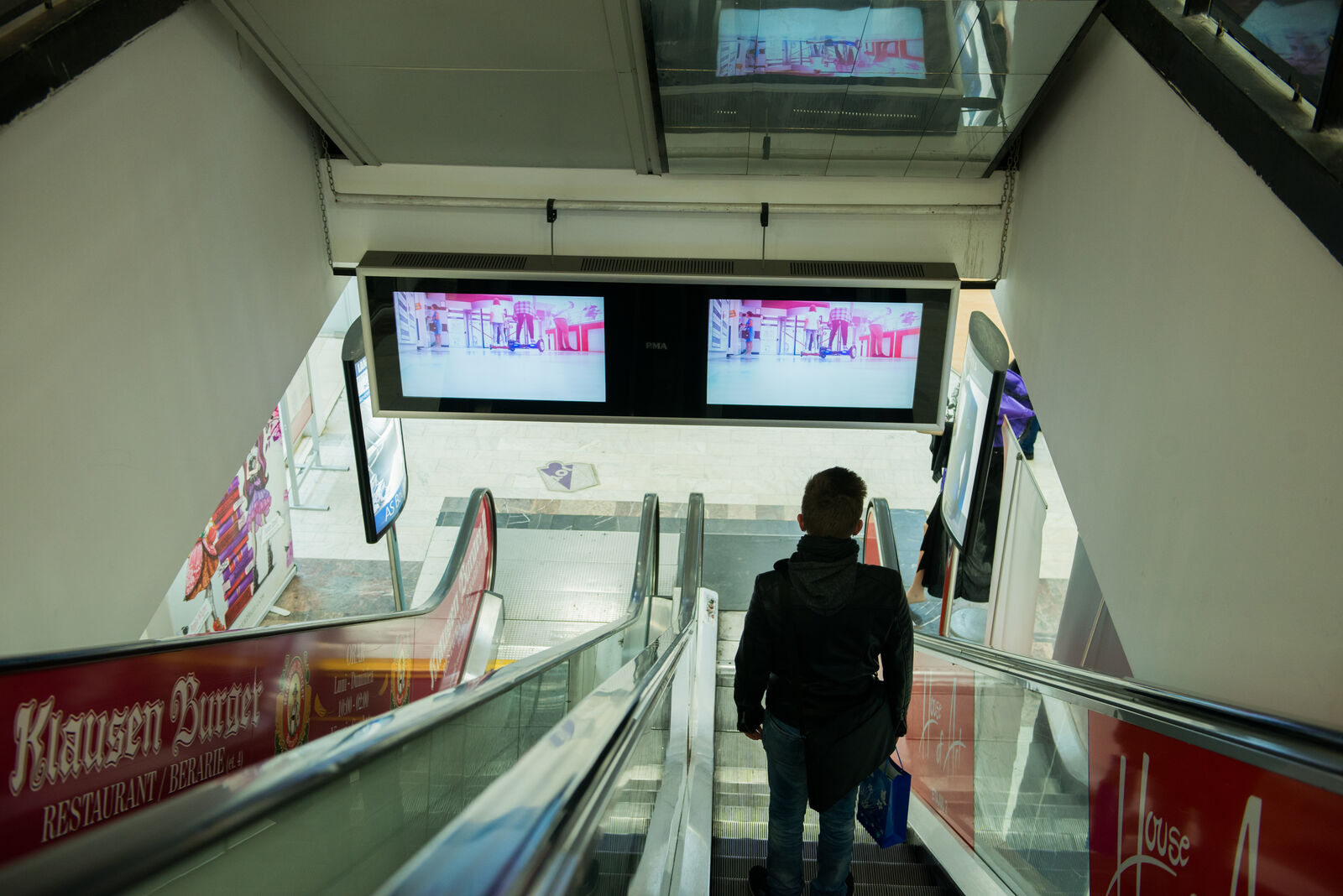 Twin landscape screens hang over a mall escalator, catching shoppers at eye level as they descend.
