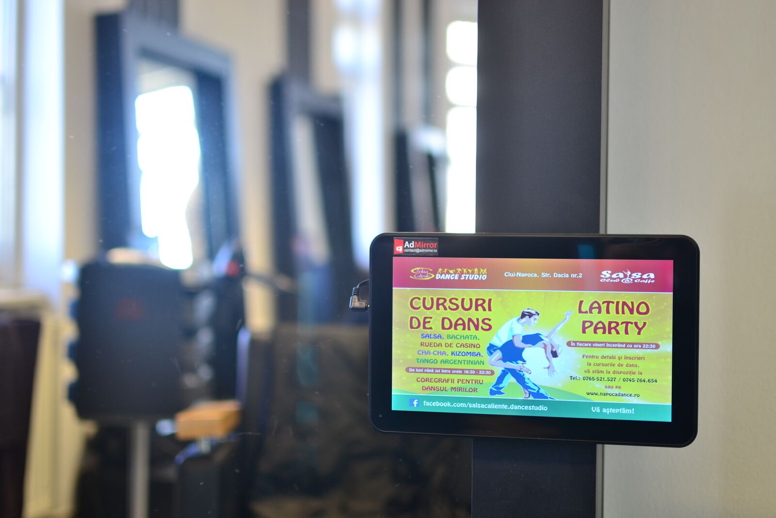 A tablet mounted on a hair-salon mirror pillar plays a bright dance-school advert; salon stations blurred behind.