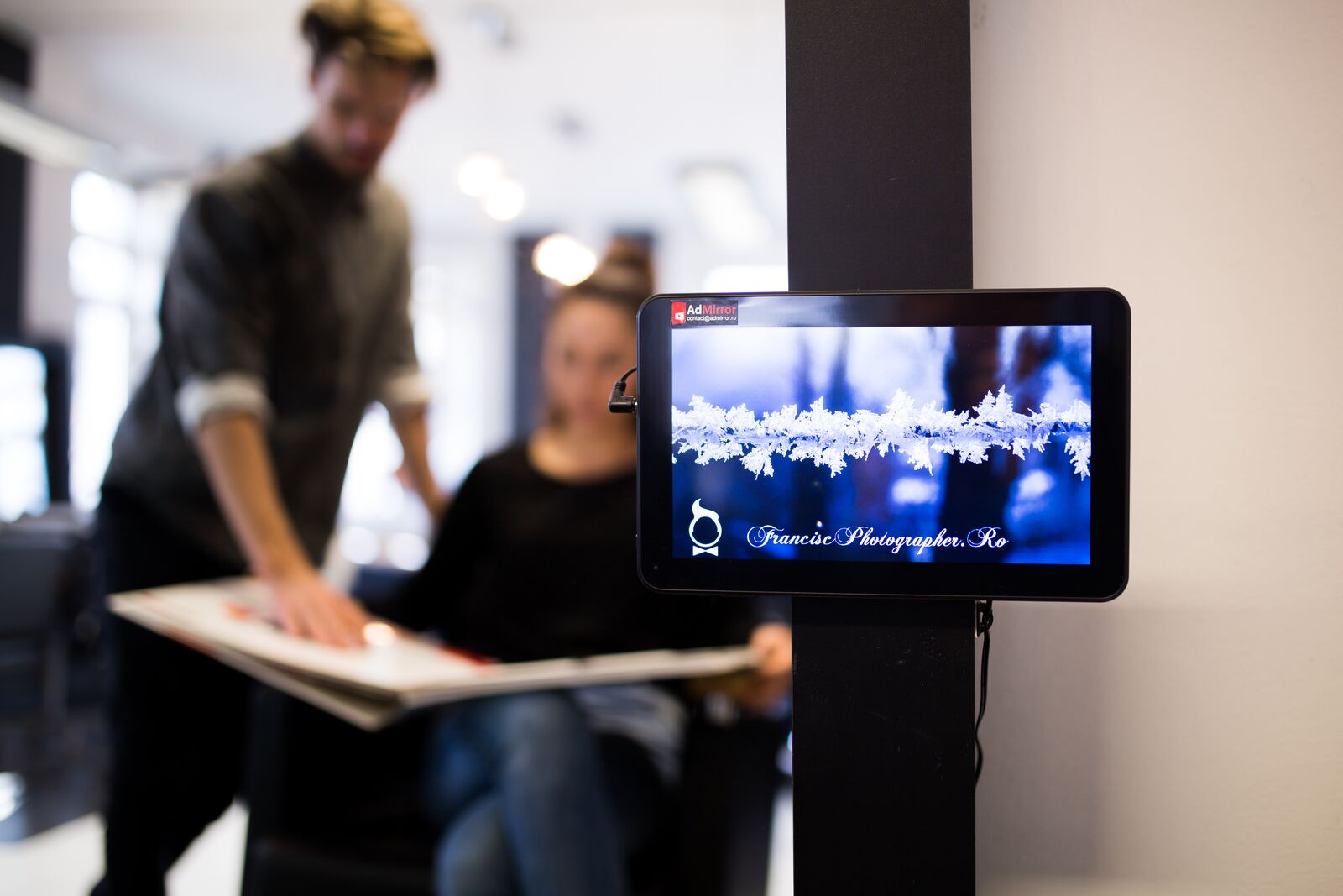 A hair-salon tablet on a mirror post plays a bright advert; a stylist and client softly out of focus behind it.