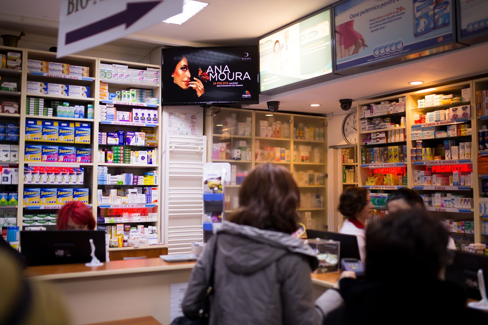 A screen above a pharmacy counter plays a mall campaign while customers wait to be served.