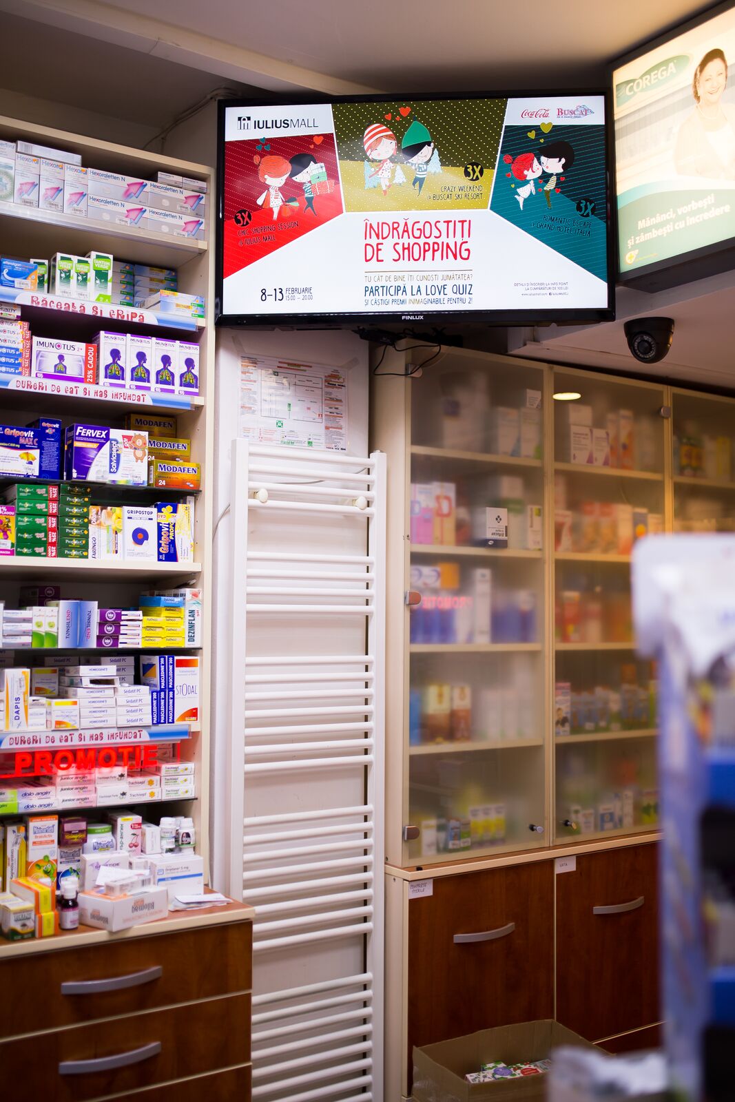 Two screens above a pharmacy counter, one carrying a shopping-mall campaign, no faces in frame.