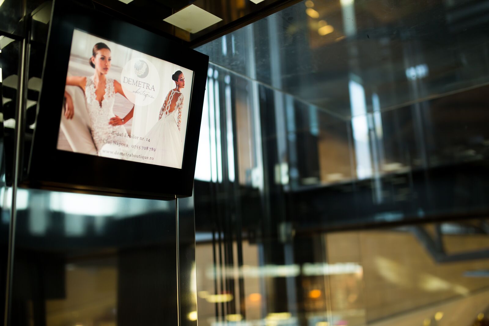 A screen fixed to the frame of a glass panoramic lift plays a bridal-boutique advert above the dark mall atrium.