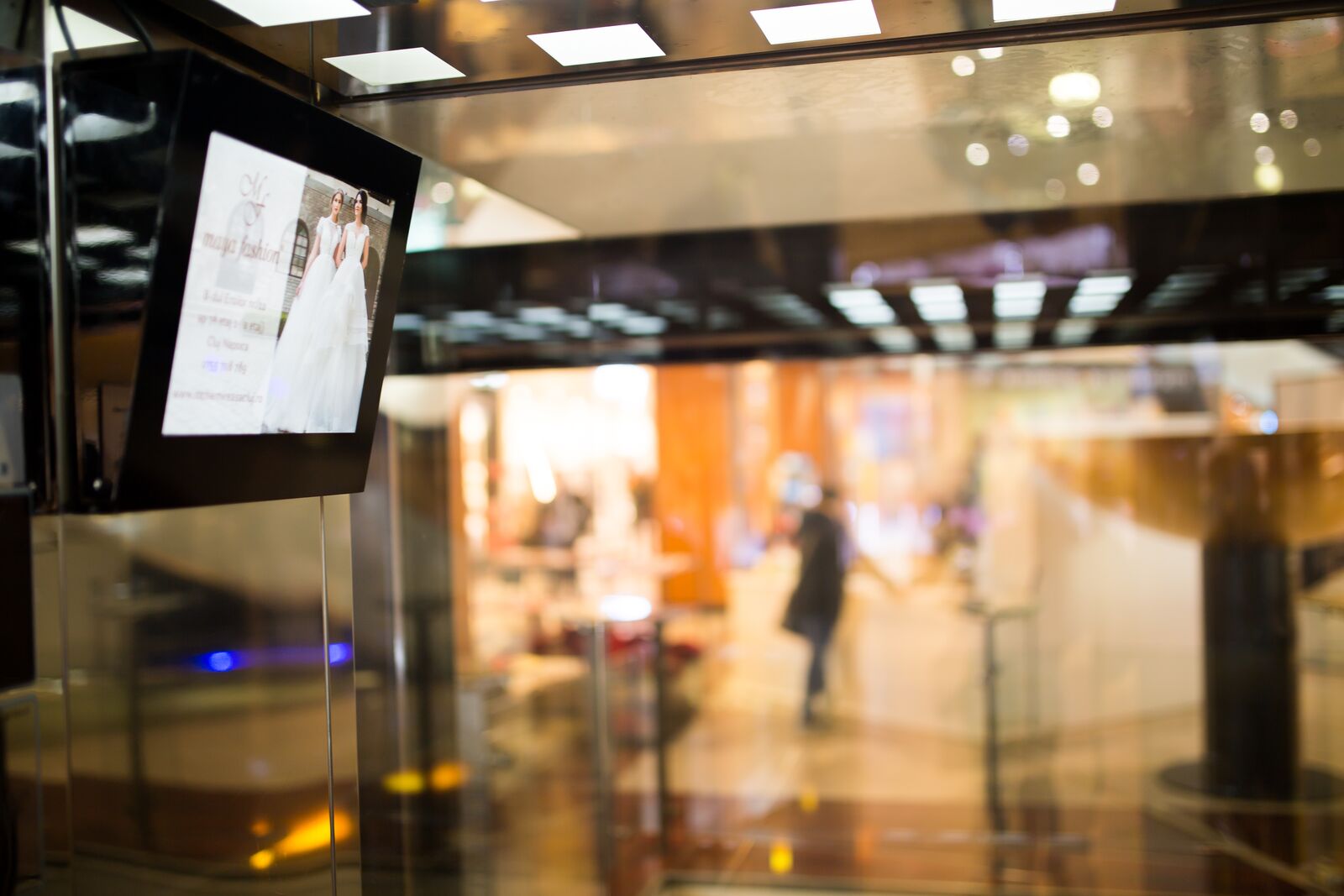 A screen at the turn of a mall walkway, warm lighting, a passer-by blurred in the middle distance.