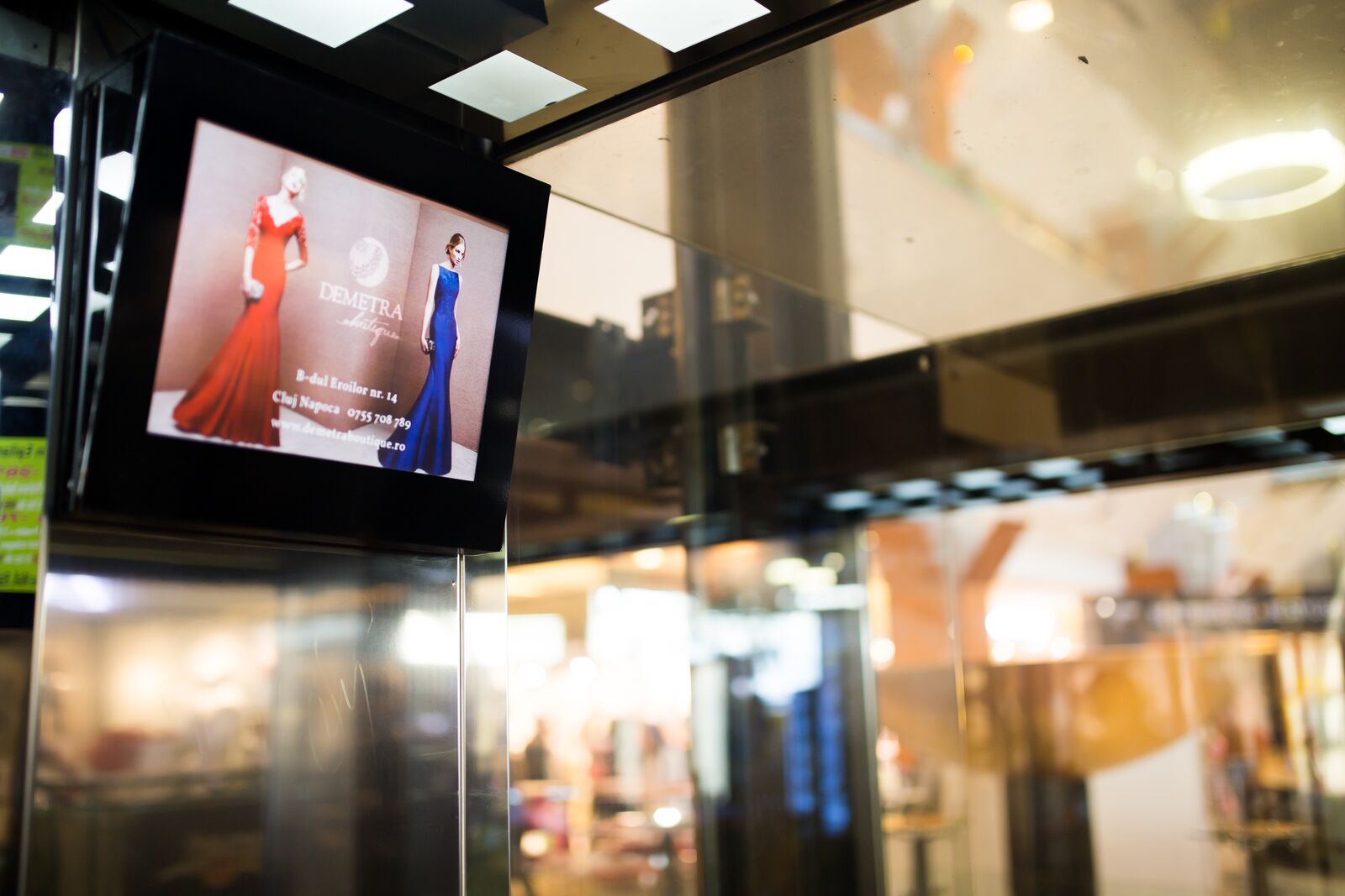 A screen on a mall lift column plays a boutique advert for two evening gowns, shopfronts out of focus behind it.