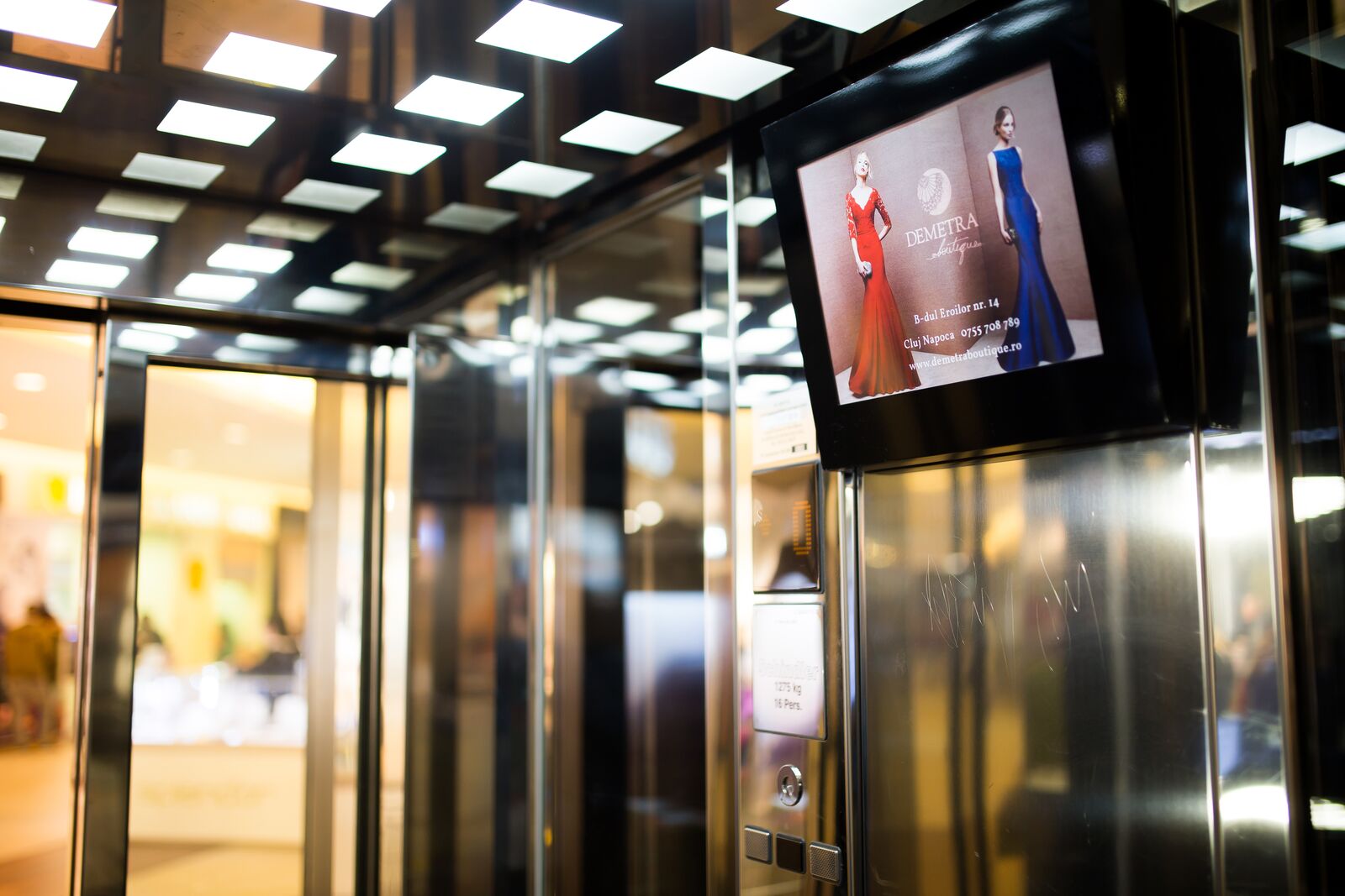 A shopping-mall lobby with a Vantano screen mounted high on the stone column beside the lift bank.