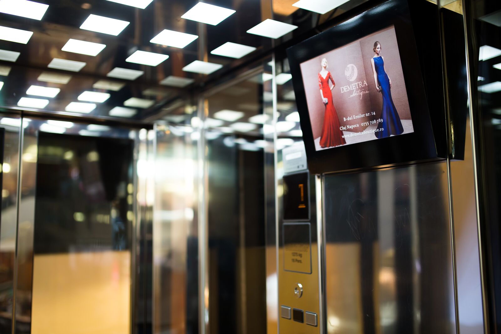 A screen mounted beside a mall lift call panel in a brass-and-glass interior under a grid of ceiling lights.